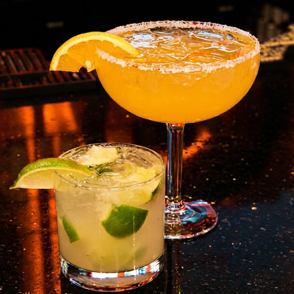 A vibrant orange margarita and a clear mojito with lime slices sit on a dark, reflective bar.