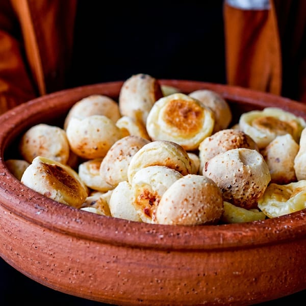 Hands holding a rustic clay bowl filled with golden-brown Brazilian cheese bread (pão de queijo).