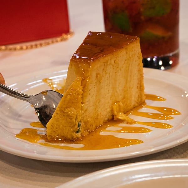 A hand with a spoon cuts into a slice of flan with caramel sauce on a white plate.
