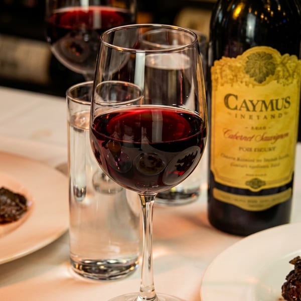 A glass of red wine, a bottle of Caymus Cabernet Sauvignon, and plates of food on a white tablecloth.